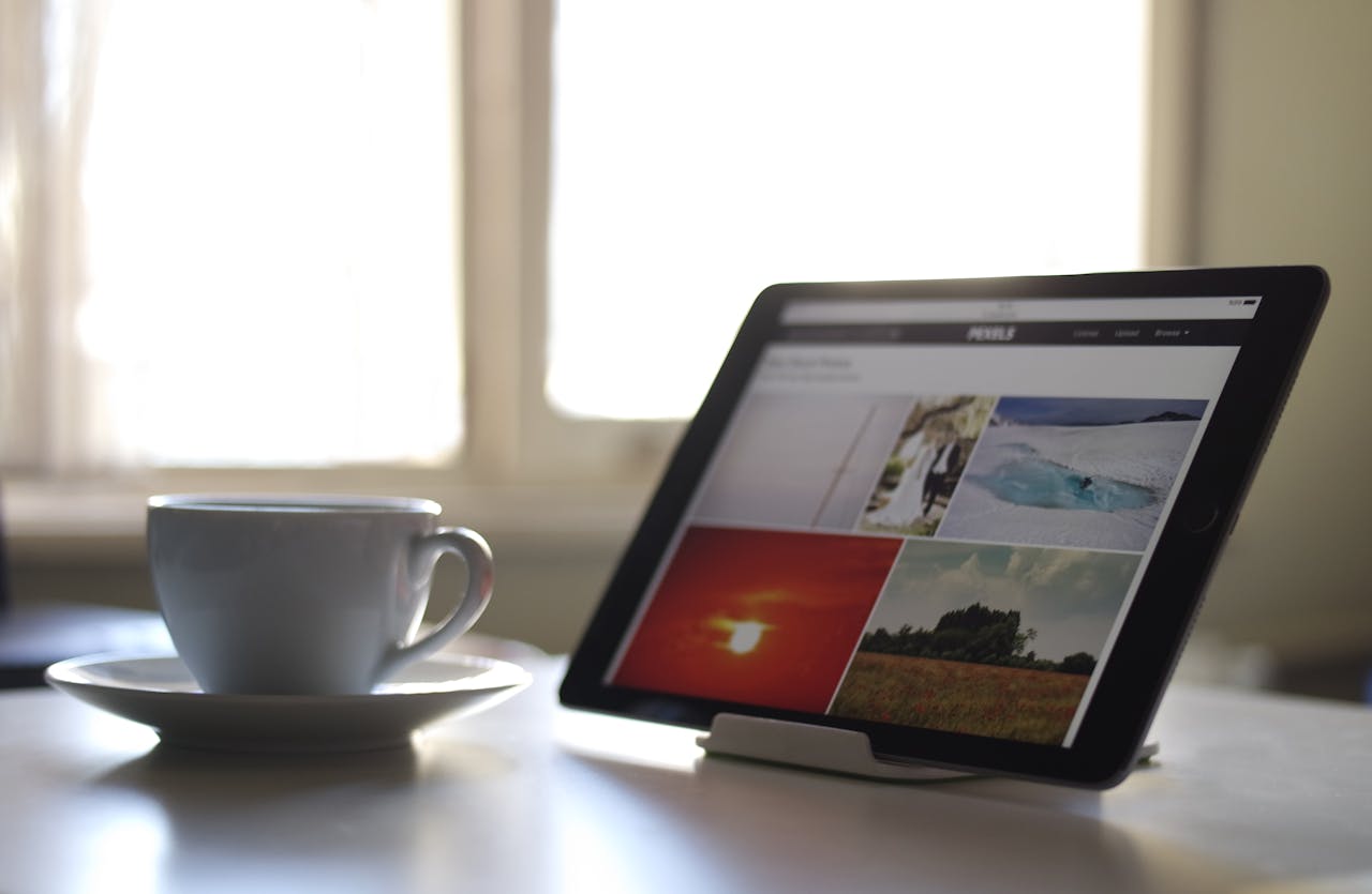 A tablet stands on a desk beside a cup of coffee, with soft daylight streaming through a window.